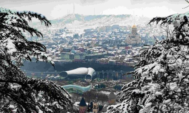 A winter city view of Tbilisi with snow on the roofs, trees, and distant mountains under a grey sky.