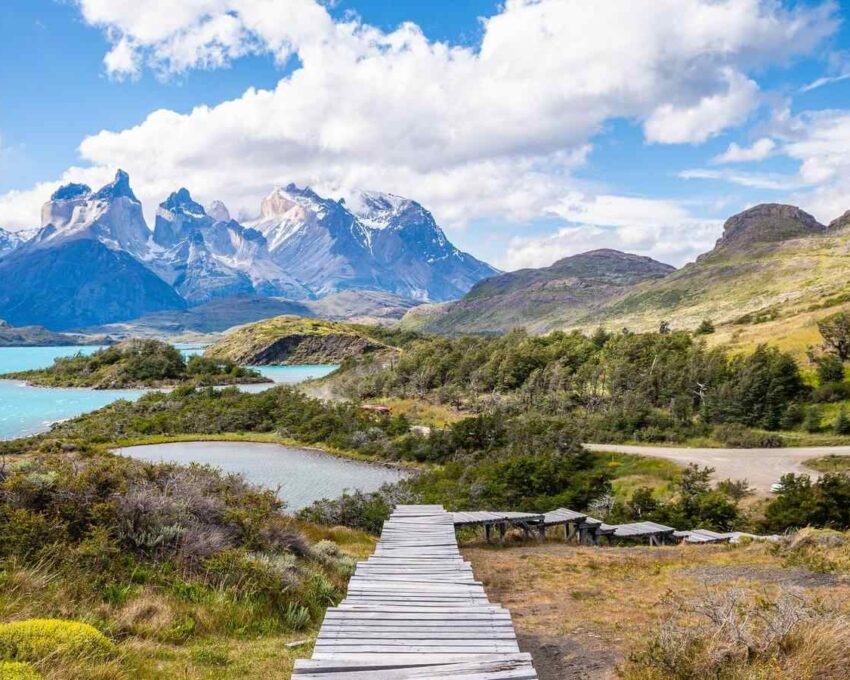 Wooden plank path leading through a valley toward bright blue lakes and steep, snow-dusted granite mountains.