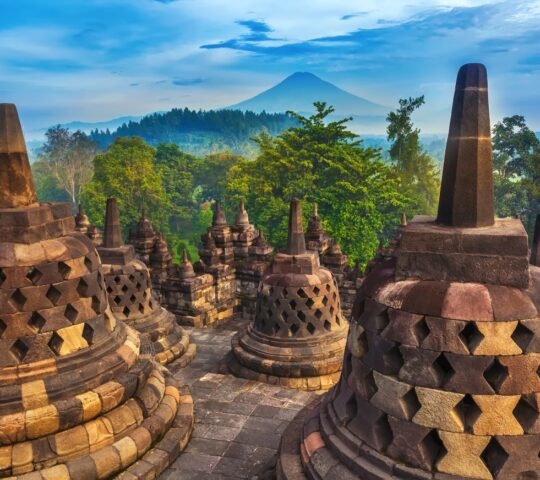 Ancient stone stupas at Borobudur Temple with a large volcanic peak and green forest in the distance.