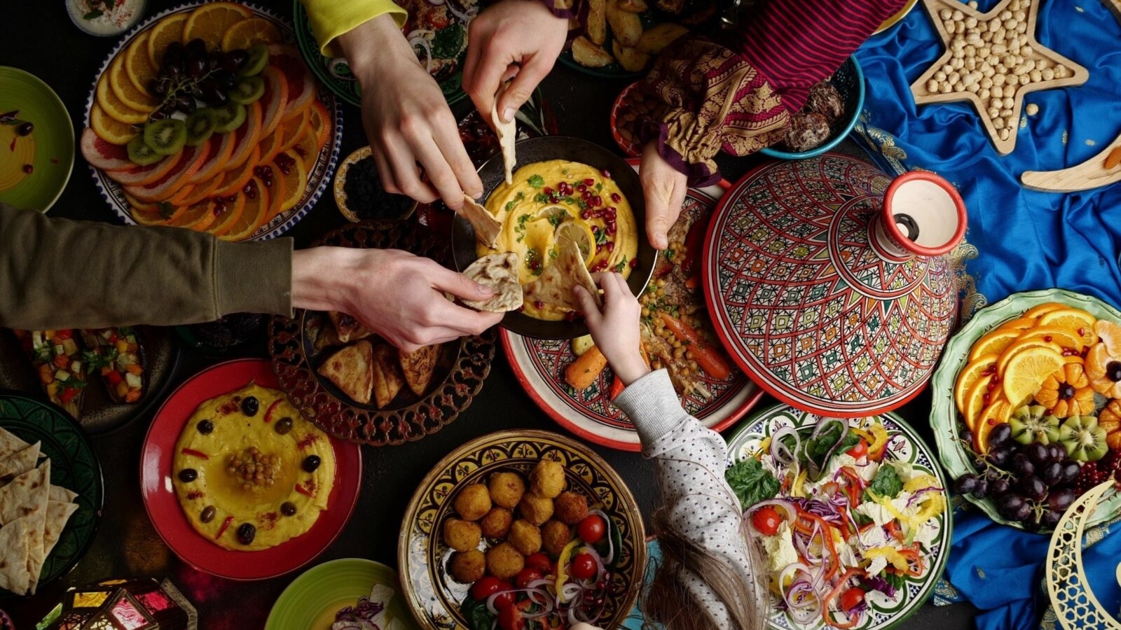 Multiple hands reaching for pita bread and hummus amidst a colorful spread of Middle Eastern dishes and fruit platters.