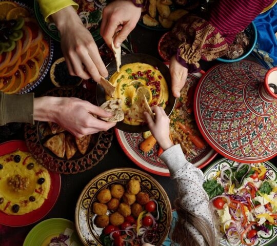 Multiple hands reaching for pita bread and hummus amidst a colorful spread of Middle Eastern dishes and fruit platters.