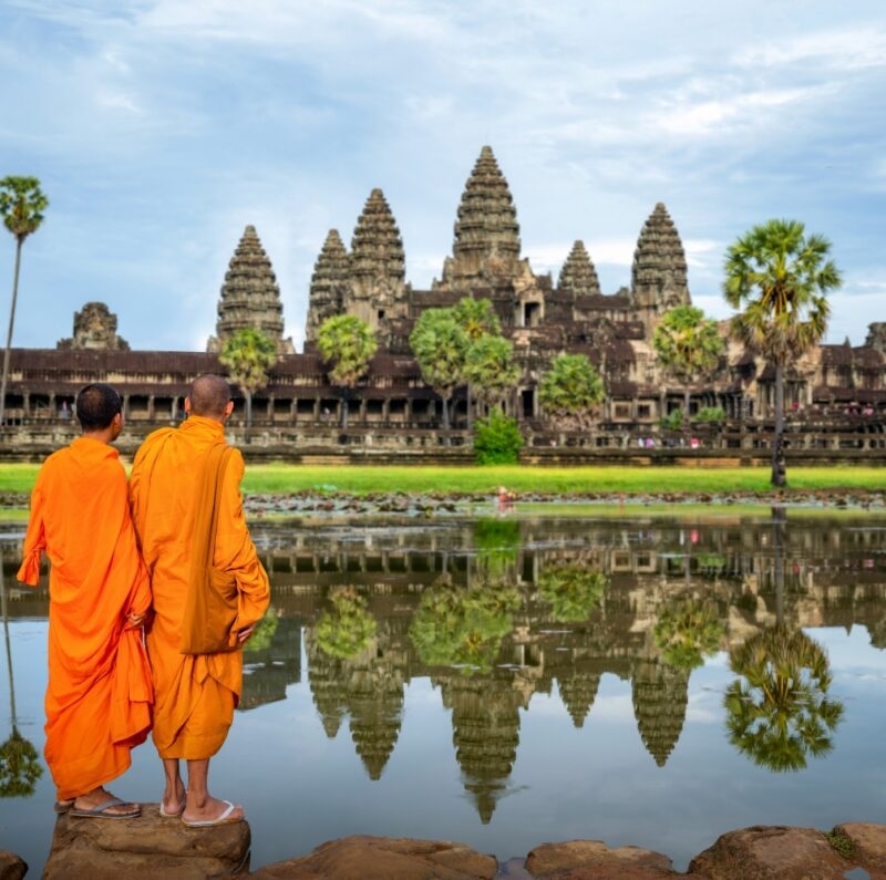 Two monks in saffron robes view the Angkor Wat temple reflected in water during luxury Asia grand tour holidays.
