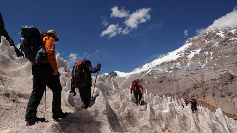 Several hikers with backpacks climbing a steep, icy slope toward a high, snow-capped mountain peak. Luxury Argentina trips.