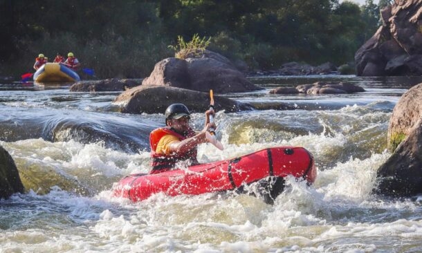 A person wearing a helmet and life vest paddles a red raft through turbulent whitewater rapids.