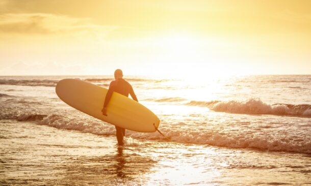 A surfer carrying a surfboard walks towards the breaking waves during a golden sunset at the beach.