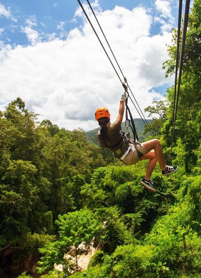 A person ziplining through a dense green tropical forest under a blue sky with white clouds.