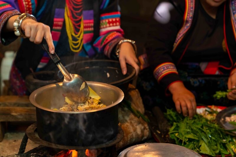 Close-up of hands stirring food in a metal pot on a stove, with people in colorful traditional ethnic dress.