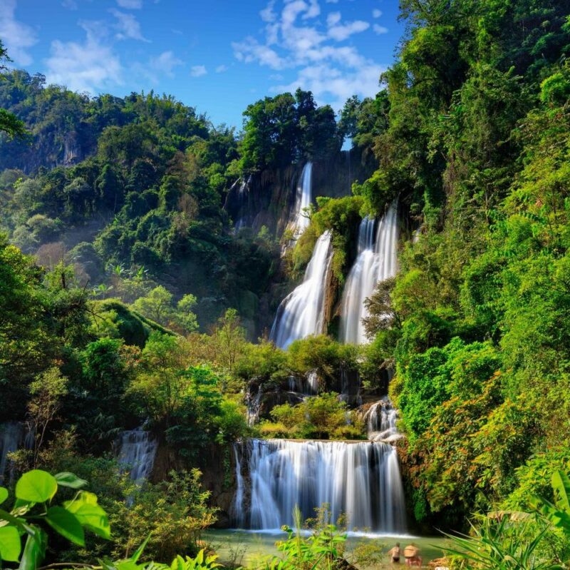 Large multi-tiered waterfall cascading down a lush green mountain into a pool surrounded by thick tropical jungle foliage.