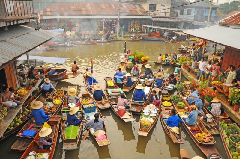 Overhead view of many small wooden boats carrying produce and vendors navigating a narrow waterway at a floating market.