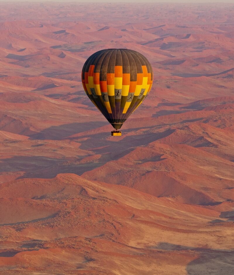 A black, orange, and yellow hot air balloon flying over a wide landscape of undulating red desert sand dunes.