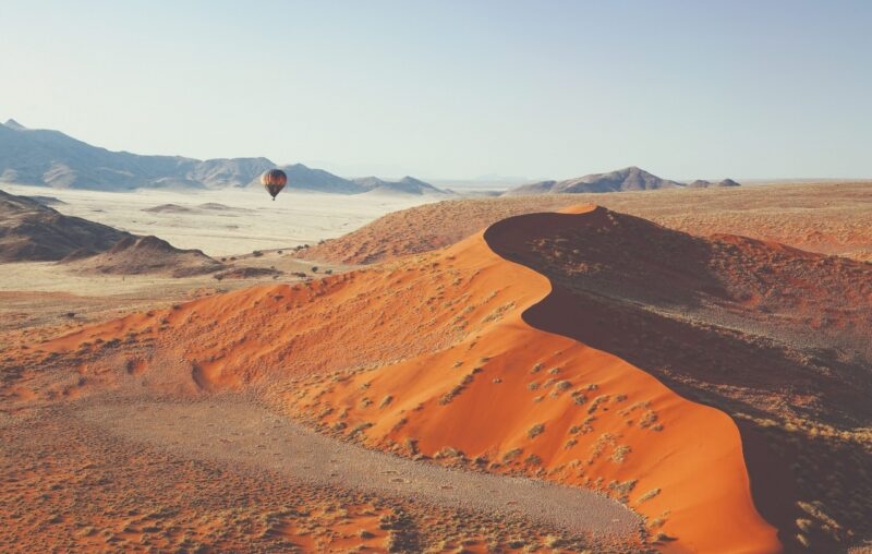Large orange sand dune in a desert valley with a hot air balloon floating in the distance under a clear sky.