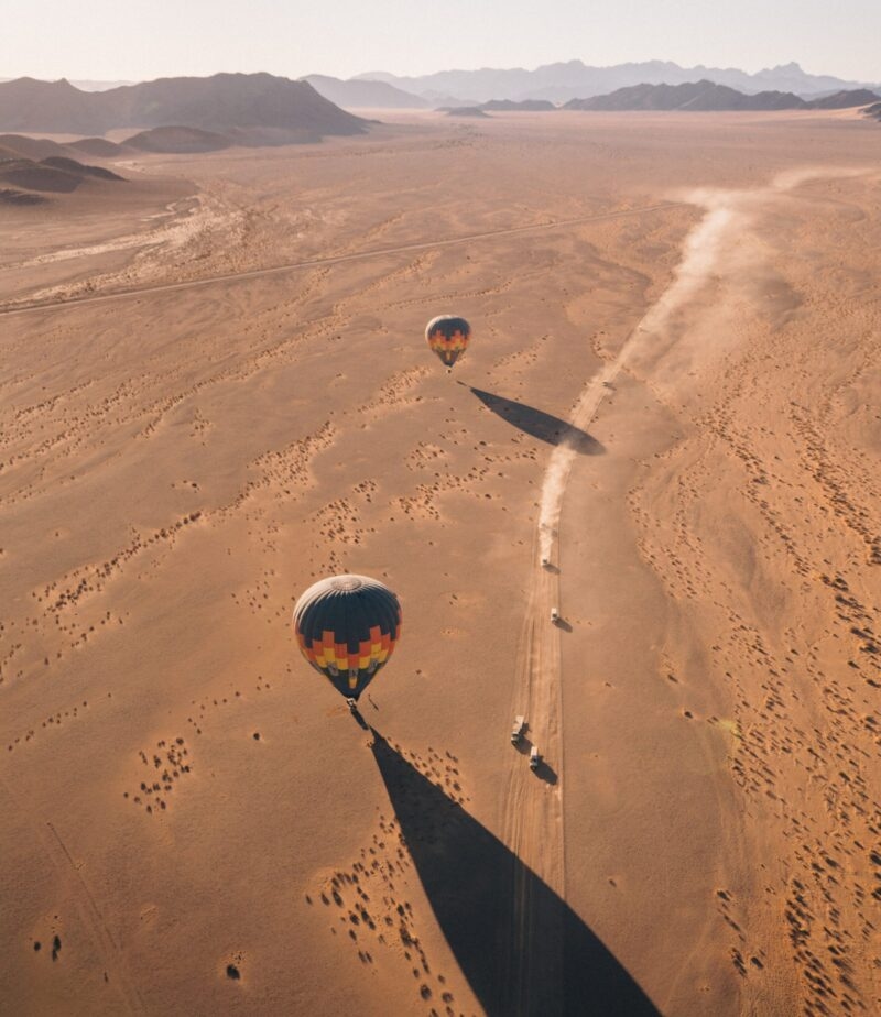 Aerial view of hot air balloons and vehicles on a dirt track in a vast desert landscape.