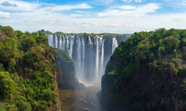 A view of the large waterfall Victoria Falls on the Zambezi River in South Africa, with many streams of water cascading down a tall cliff face in to a wide river