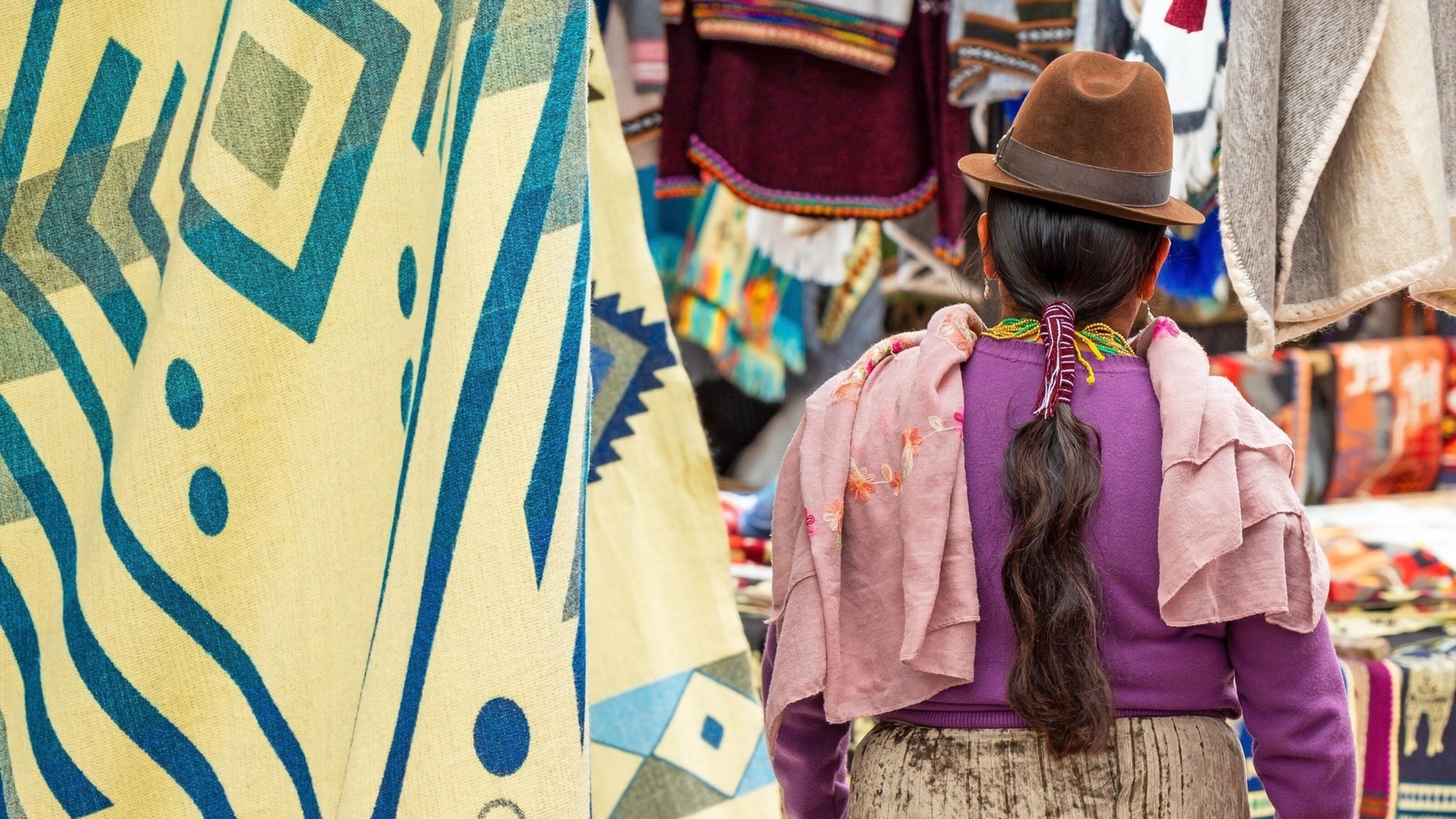 Indigenous Ecuadorian Otavalo woman in traditional clothing, hat and hairstyle on Otavalo local market with textile and fabric stalls, Ecuador.