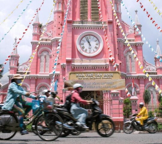 Traffic moves past a large, bright pink Gothic-style church with a clock tower.