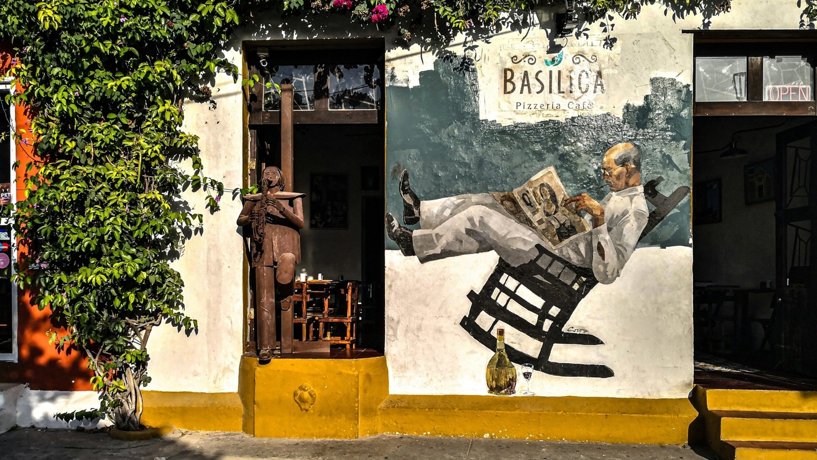 A vibrant café entrance adorned with a mural of a man relaxing in a rocking chair, framed by lush greenery and rustic decor.