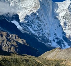 Two hikers stand in a lush valley, gazing up at a towering snow-capped mountain ridge under a clear blue sky.