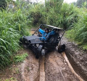 A small off-road vehicle is stuck in a muddy trail surrounded by tall, green grass and dense vegetation.