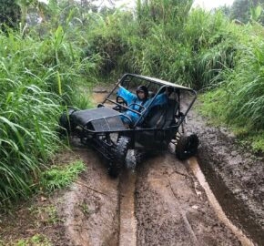 A small off-road vehicle is stuck in a muddy trail surrounded by tall, green grass and dense vegetation.