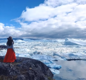 A woman in a red skirt and hat stands on rocky terrain, gazing at icebergs and a reflective sea under a cloudy blue sky.