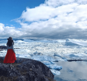 A woman in a red skirt and hat stands on rocky terrain, gazing at icebergs and a reflective sea under a cloudy blue sky.