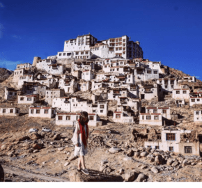 A woman gazes up at a mountainside village with traditional white buildings, set against a clear blue sky and rugged terrain.