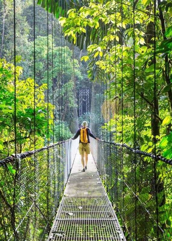 A hiker with a yellow backpack walking across a narrow suspension bridge in a lush green jungle.