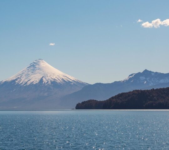 A large snow-covered volcanic peak stands behind a dark shoreline across a calm expanse of blue water.