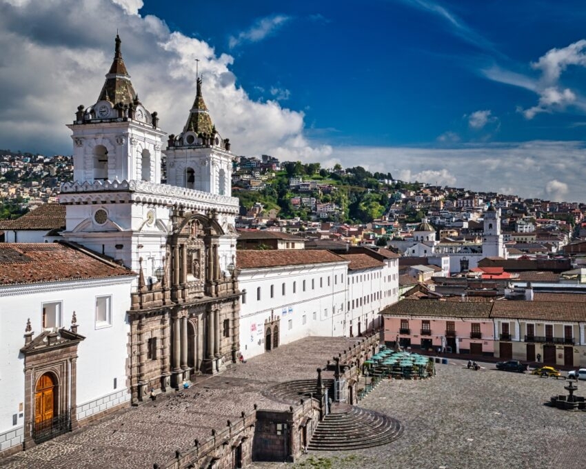 Ornate white church with twin steeples overlooking a historic square and terraced cityscape under a cloudy blue sky.