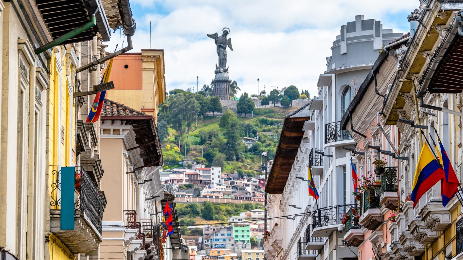 A historic street in Quito with buildings flanking both sides and the El Panecillo statue in the background.