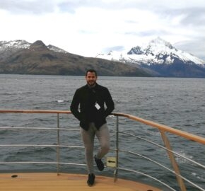 Person on a boat deck with snow-capped mountains and a lake in the background.
