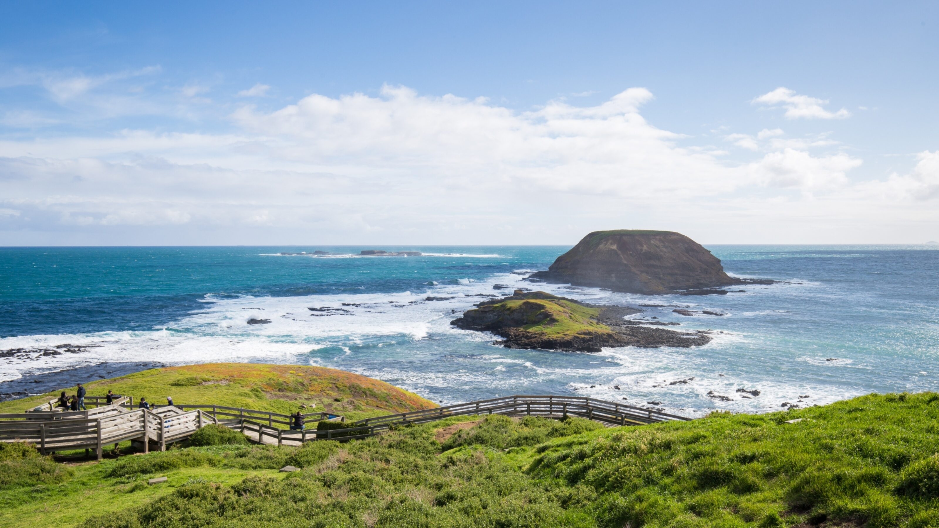 Coastal view with grassy cliffs, a wooden walkway, and a rocky island under a blue sky.