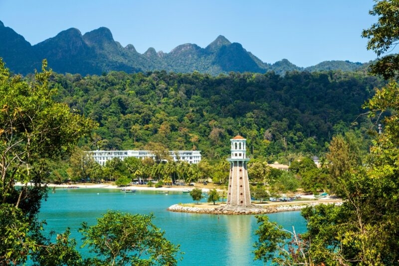 A stone lighthouse overlooks a calm blue bay surrounded by lush mountains on luxury Malaysia holidays.
