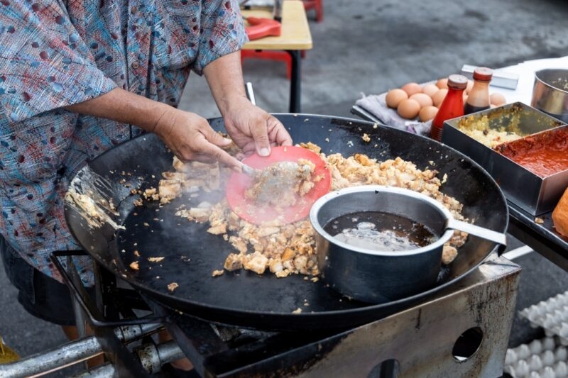 A chef stir-fries food in a large pan at a street stall during luxury Malaysia tours.