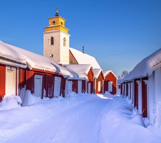 Snow-covered path flanked by red wooden houses leading to a white church with a yellow bell tower.