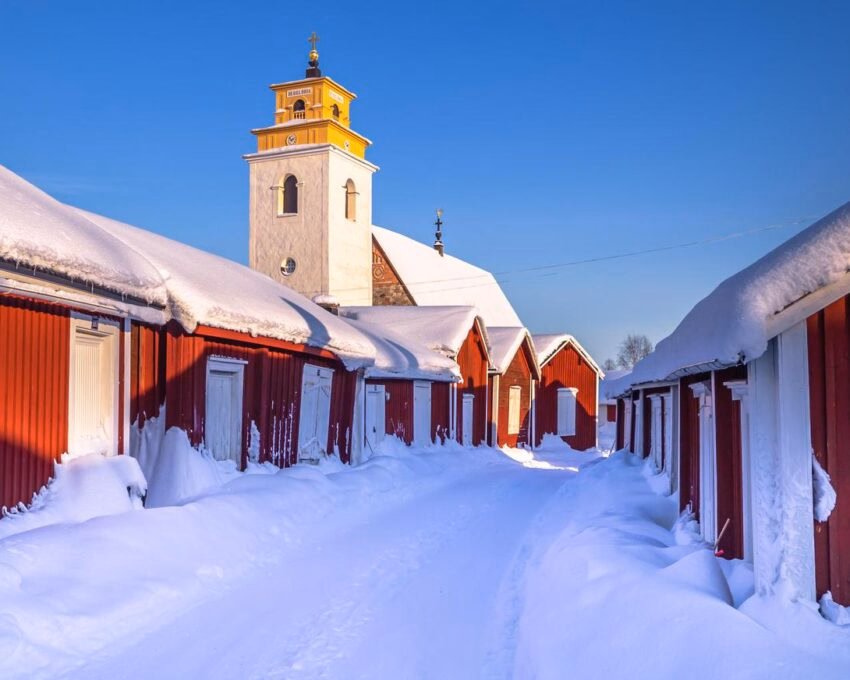 Snow-covered path flanked by red wooden houses leading to a white church with a yellow bell tower.