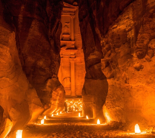 A warmly lit narrow canyon leading to an ancient, carved facade by candlelight.