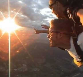 Skydiver in harness reaching out towards the sunset mid-air.