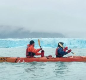 Two people kayaking near a glacier with overcast skies.