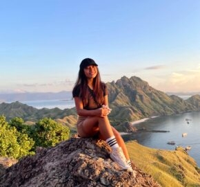 Person sitting atop a rocky hill with scenic views of mountains and sea at sunset.