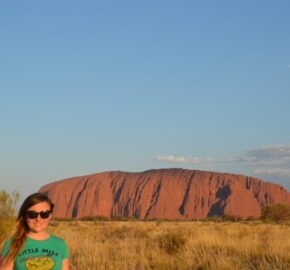 Person standing in front of Uluru at sunset with blue sky and sparse clouds above.