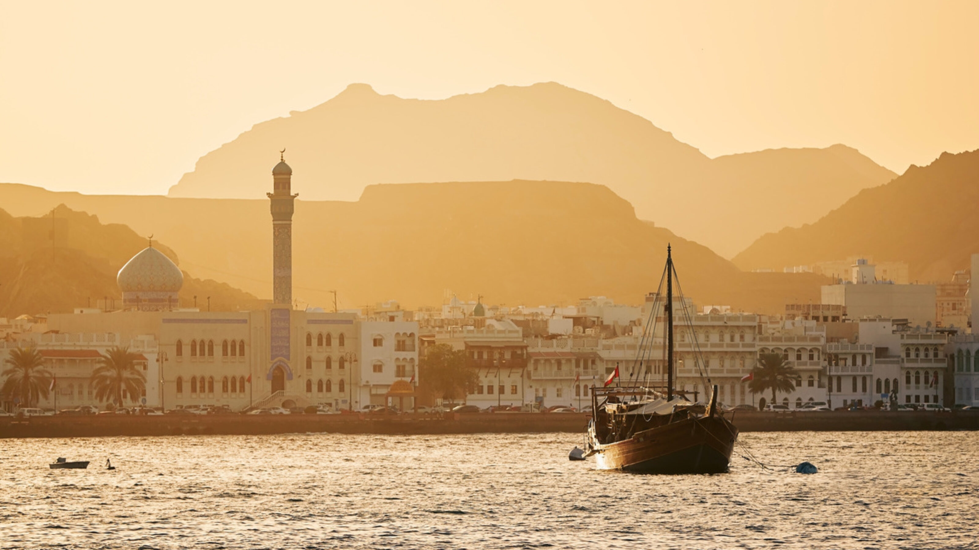 A traditional Dhow floating in the water with Muscat Old Town in the background
