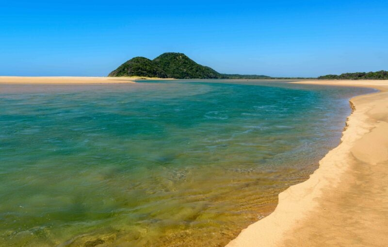 Tranquil beach with clear turquoise water, golden sand, and a lush green hill under blue sky.