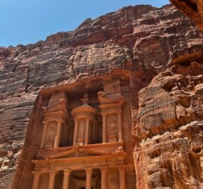 View of the ancient facade of Al-Khazneh carved into red cliffs under a clear blue sky in Petra, Jordan.