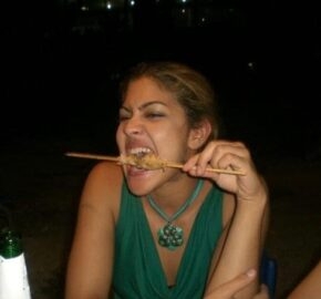 Person in a green top eating chicken off of a wooden skewer, with a bottle on the table.