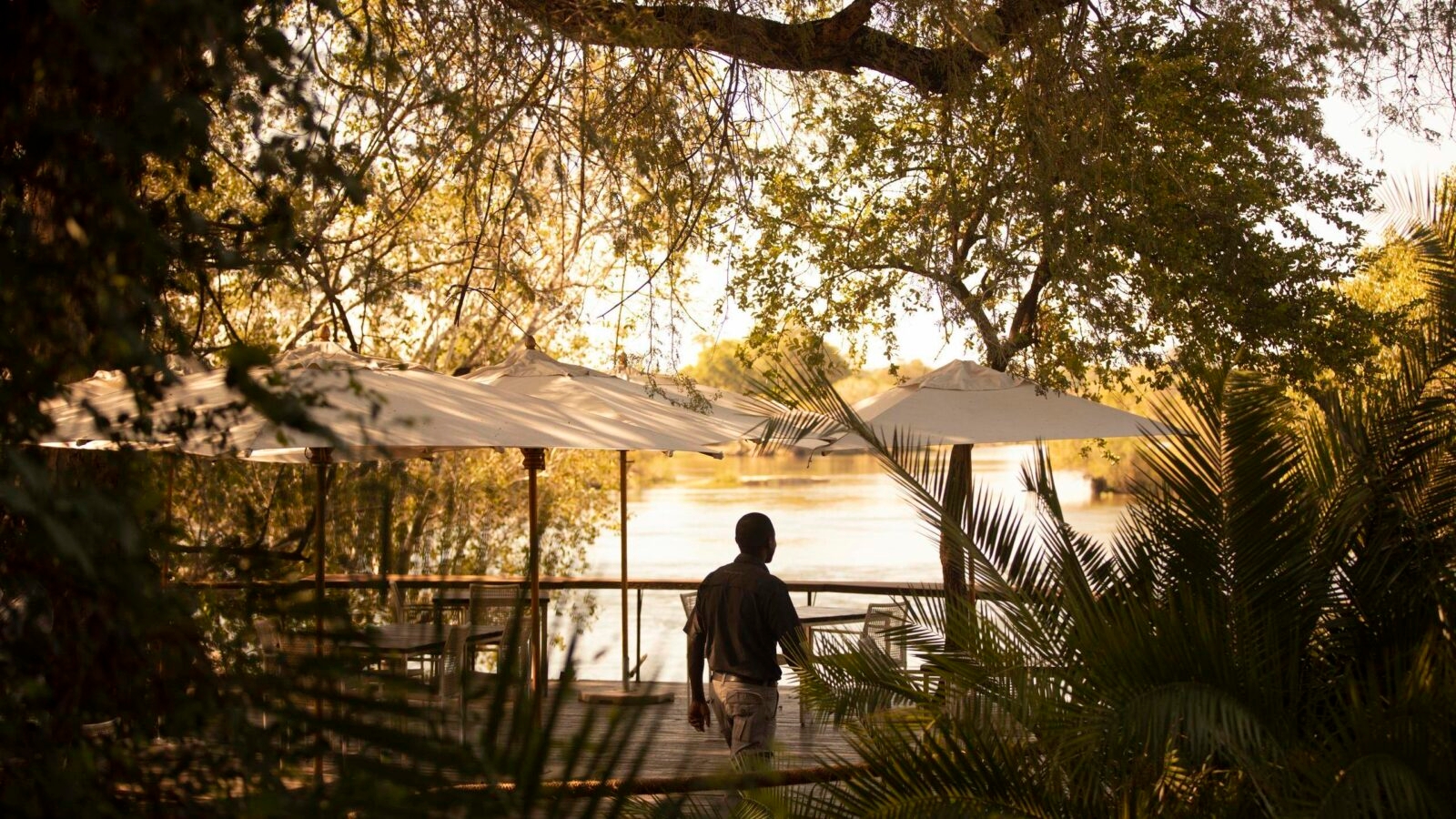 Person on a deck with umbrellas overlooking a tranquil river at sunset, surrounded by trees.