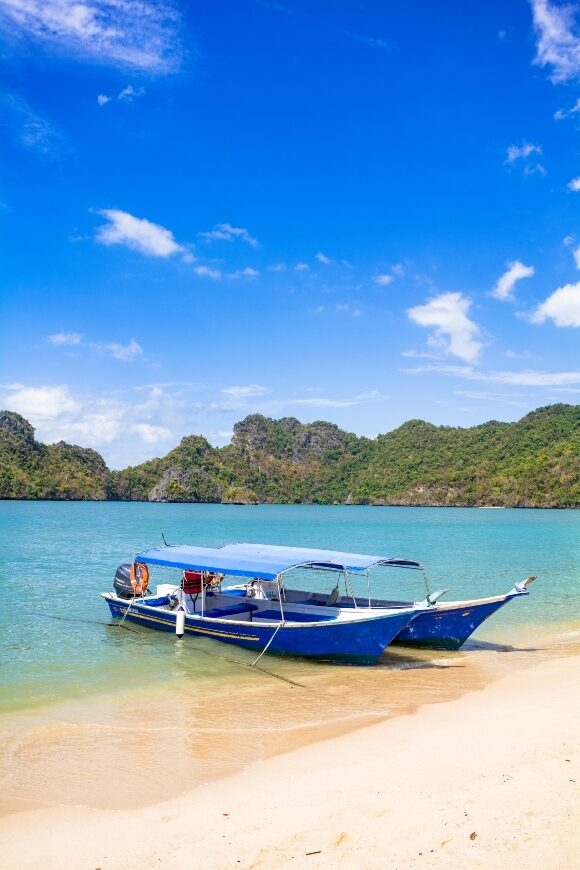 A blue and white boat moored on a sunny tropical beach with clear water and green hills.
