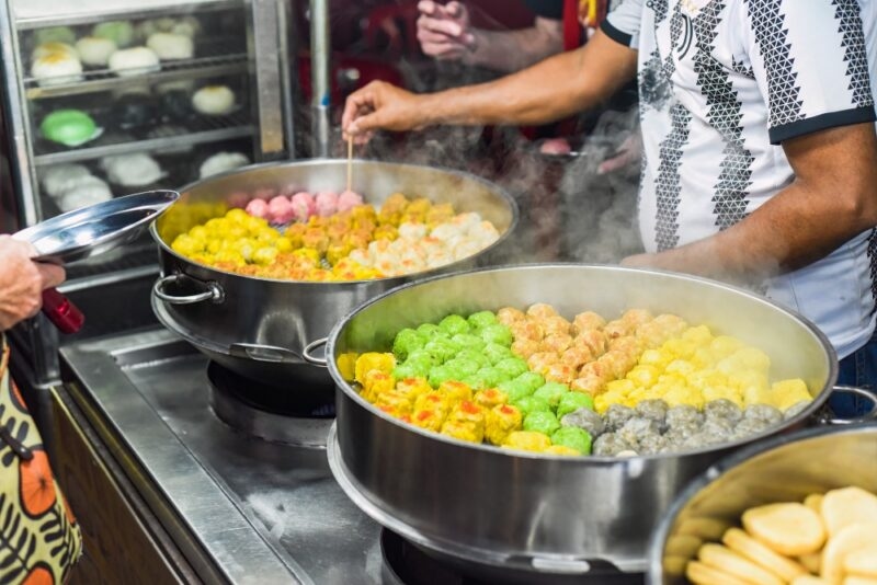Colorful dumplings in steaming pots at a street food stall, with vendors preparing food.
