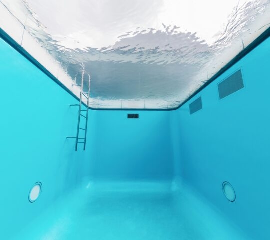 Perspective view from inside an empty blue pool looking up at a rippling water surface and metal ladder.