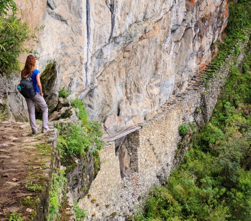 Young woman enjoying the view of Inca Bridge and cliff path near Machu Picchu in Peru. The Bridge is a part of a mountain trail that heads west from Machu Picchu